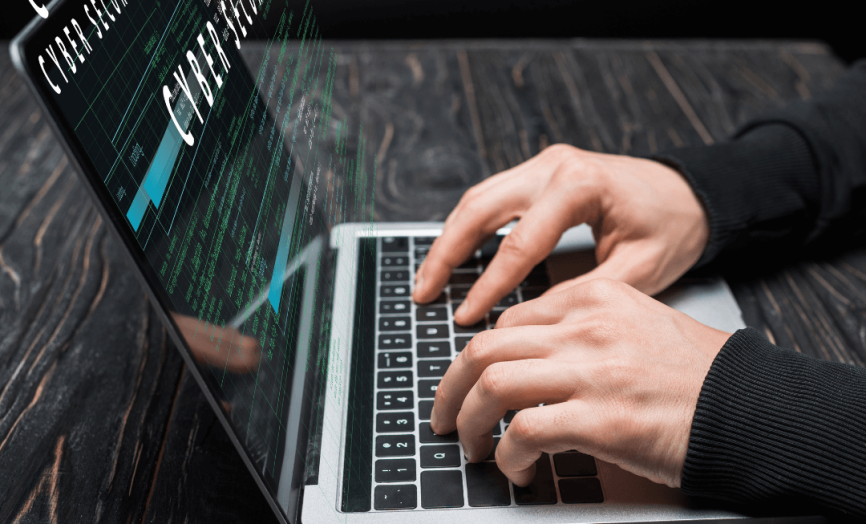 Detailní pohled na ruce píšící na notebooku, ze kterého vystupují holografické nápisy „CYBER SECURITY“ na tmavém dřevěném pozadí.