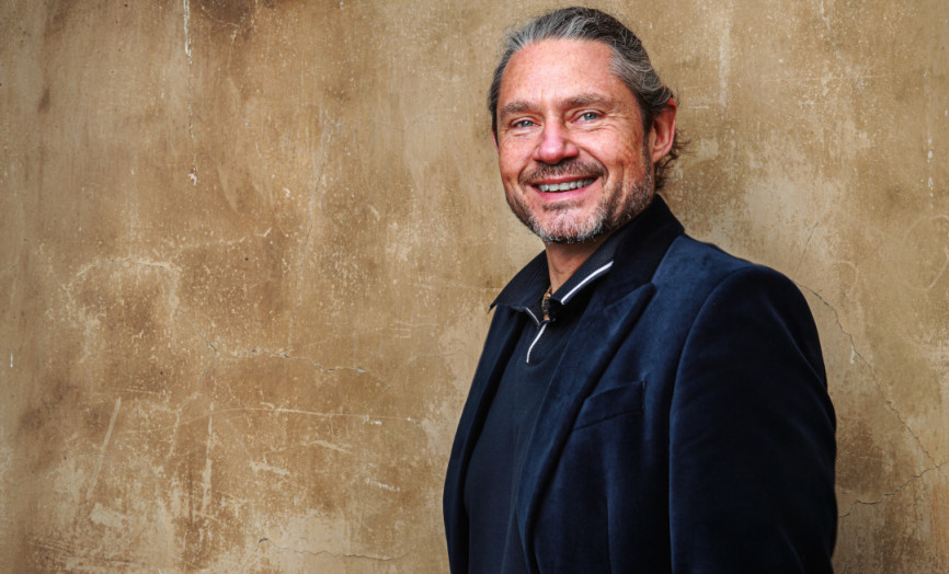 Osoba stojí před hnědou, opotřebovanou stěnou s popraskanou texturou, oblečená v tmavém saku a tmavé košili se zipem, s úsměvem hledící do kamery