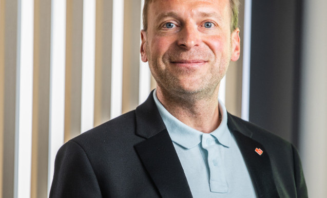 Osoba stojí v interiéru před stěnou z vertikálních dřevěných lamel, oblečená v černém saku a světle modrém polo triku s červeným odznakem na klopě