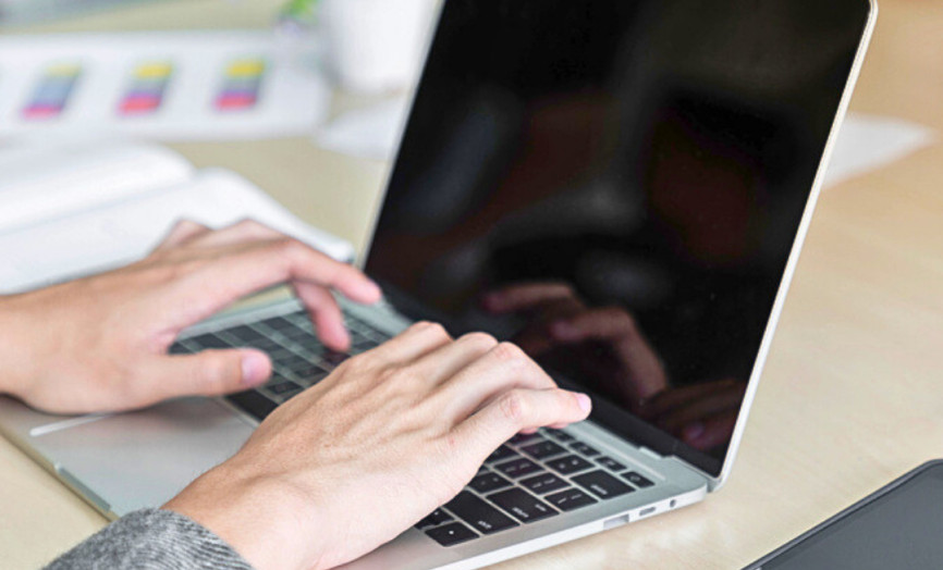 Osoba píše na notebooku u stolu s tabletem, hrnkem, zápisníkem a malou rostlinou v moderním pracovním prostředí