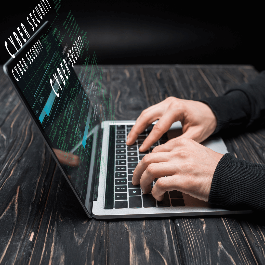 Detailní pohled na ruce píšící na notebooku, ze kterého vystupují holografické nápisy „CYBER SECURITY“ na tmavém dřevěném pozadí.
