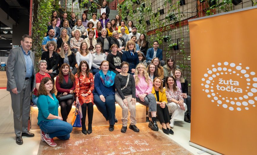 Velká skupina lidí, převážně žen, pózuje na vnitřním schodišti obklopeném vertikální zahradou s popínavými rostlinami; v popředí stojí vpravo výrazný oranžový roll-up banner s bílým logem a nápisem „žuta točka“.