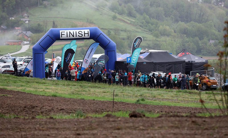 Pohled na modrou nafukovací bránu s nápisem „FINISH“ uprostřed kopcovité krajiny; pod bránou a v jejím okolí se shromažďuje velký počet účastníků v barevném sportovním oblečení, v pozadí jsou vidět týmové stany a zaparkovaná auta pod zamračenou oblohou.