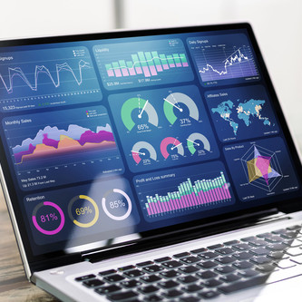 Osoba sedí před notebookem a v ruce drží chytrý telefon, oba displeje zobrazují barevné datové vizualizace – grafy, mapy a ukazatele výkonu – působí to jako práce s analytickým nebo business‑intelligence dashboardem