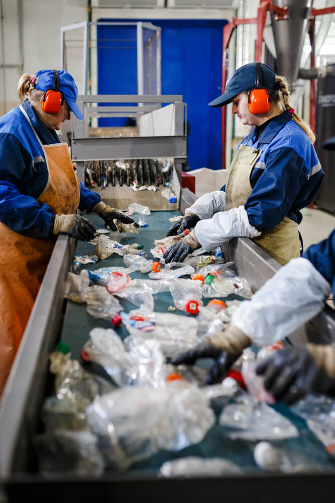 Ilustrační vizuál s pracovníky třídícími plastový odpad na pásu v recyklačním provozu – ochranné pomůcky, drcené lahve a průmyslové prostředí