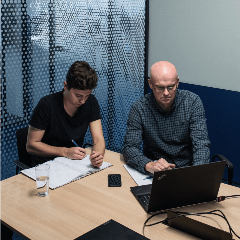 Dva lidé při pracovním jednání v zasedací místnosti, jeden píše poznámky do sešitu, druhý pracuje na notebooku