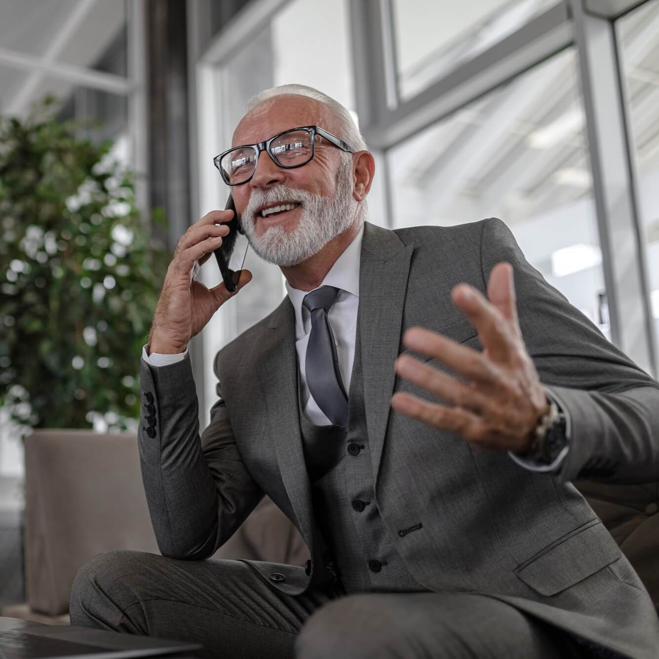 Obchodné prostredie: starší muž v sivom obleku telefonuje a gestikuluje v modernej kancelárii s rastlinou