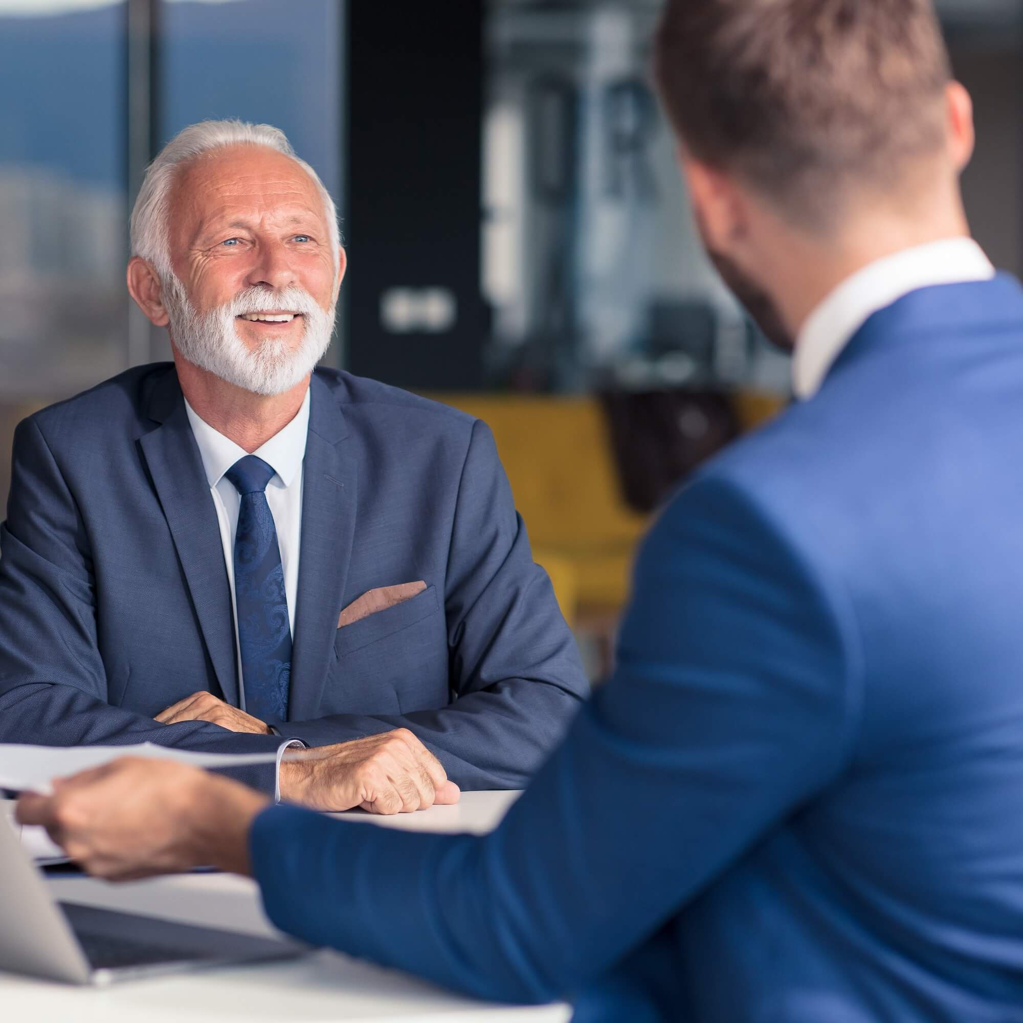 Obchodné stretnutie: dvaja muži v oblekoch pri stole, jeden drží dokument, druhý sa usmieva a počúva