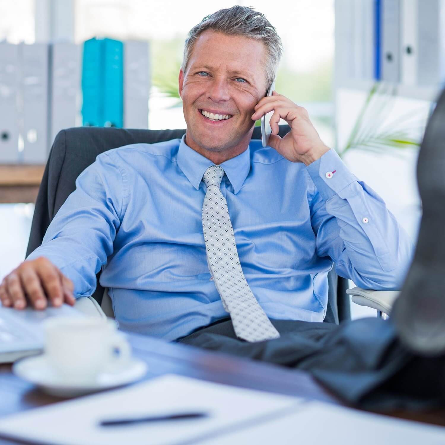 Pracovní telefonát: muž v košili a kravatě telefonuje u stolu s notebookem, šálkem kávy a dokumenty