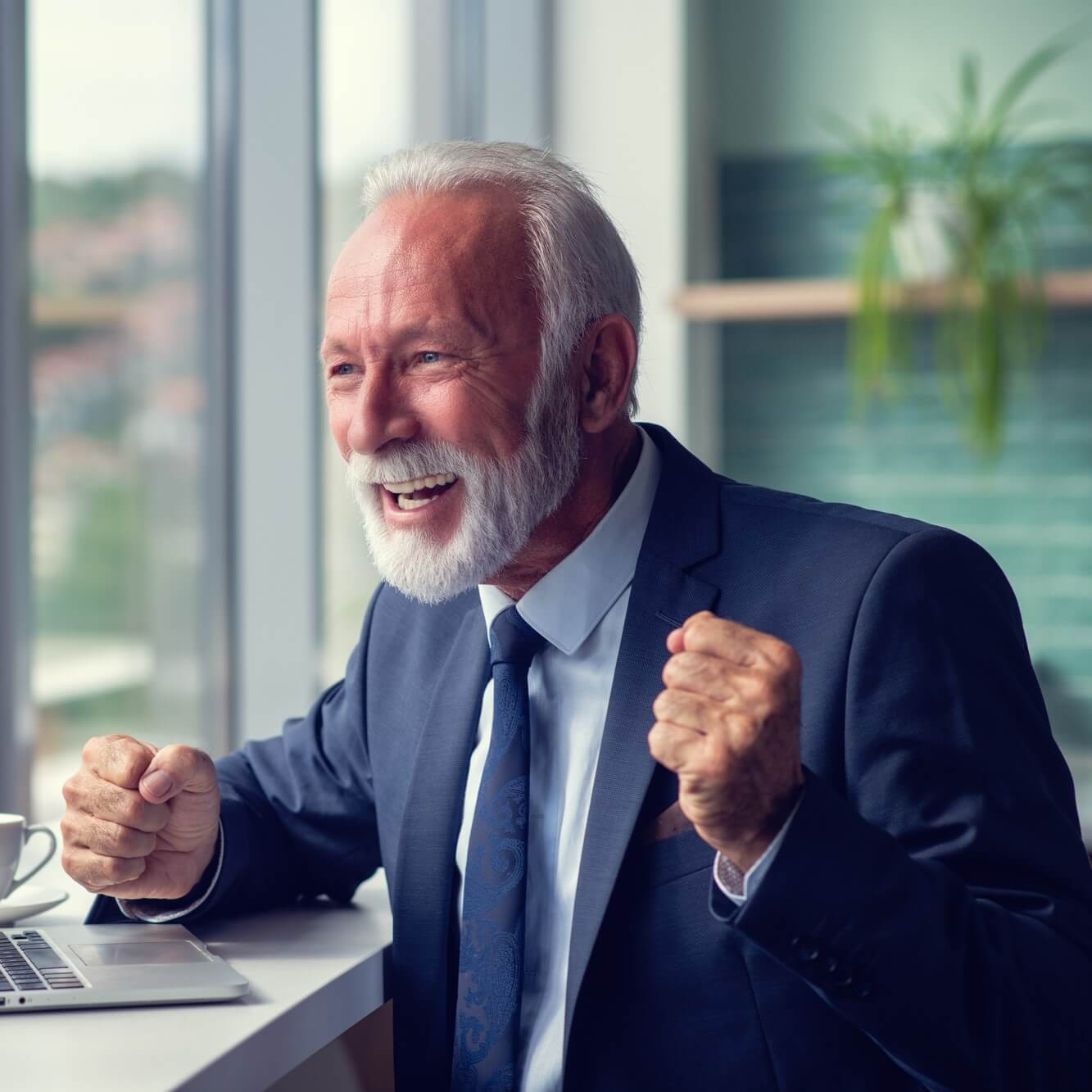 Pracovný úspech: muž v obleku pri notebooku s úsmevom a víťazným gestom, šálka kávy na stole