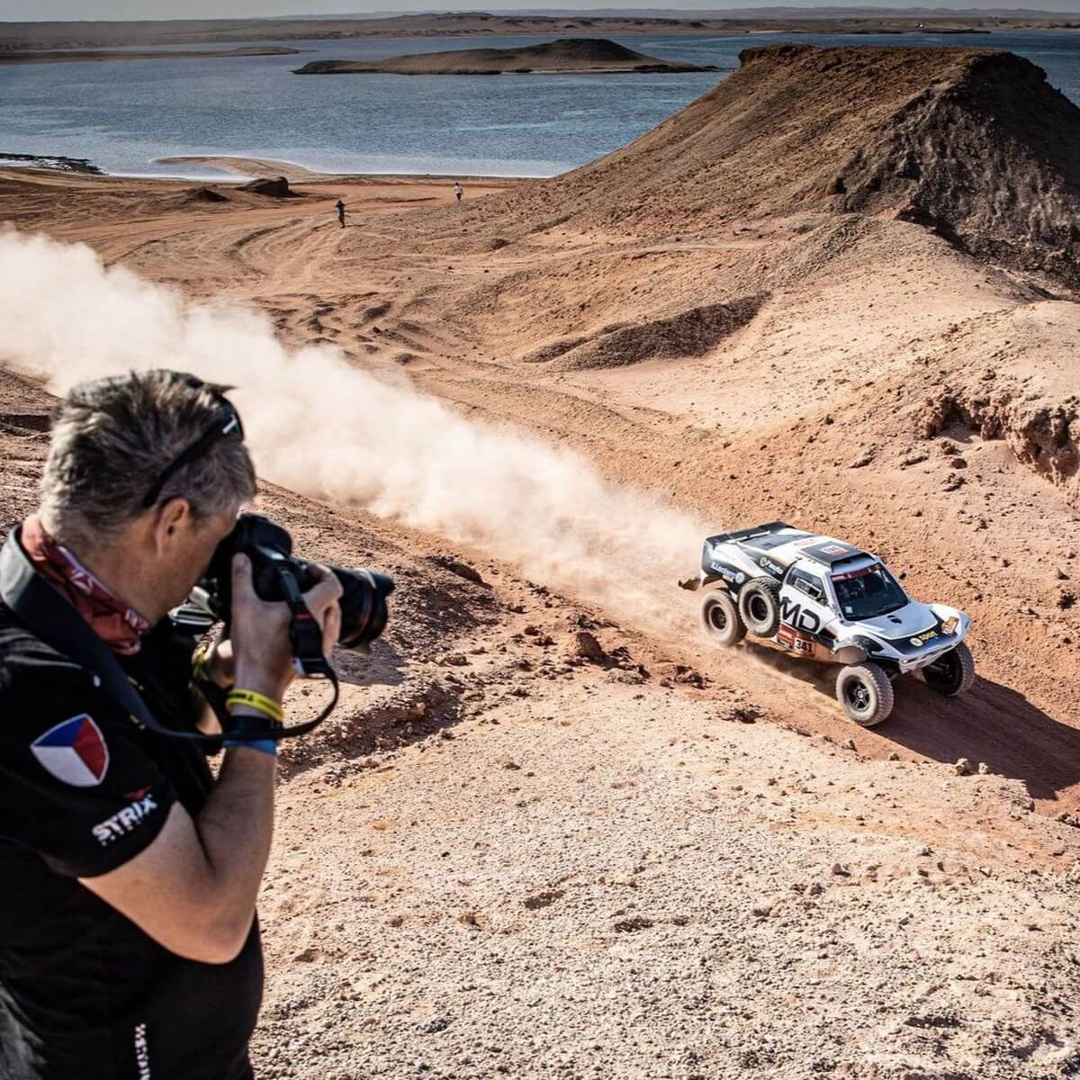 Závodní truck č. 321 s logem MD projíždí prašnou pouštní krajinou u vodní plochy, fotograf v popředí