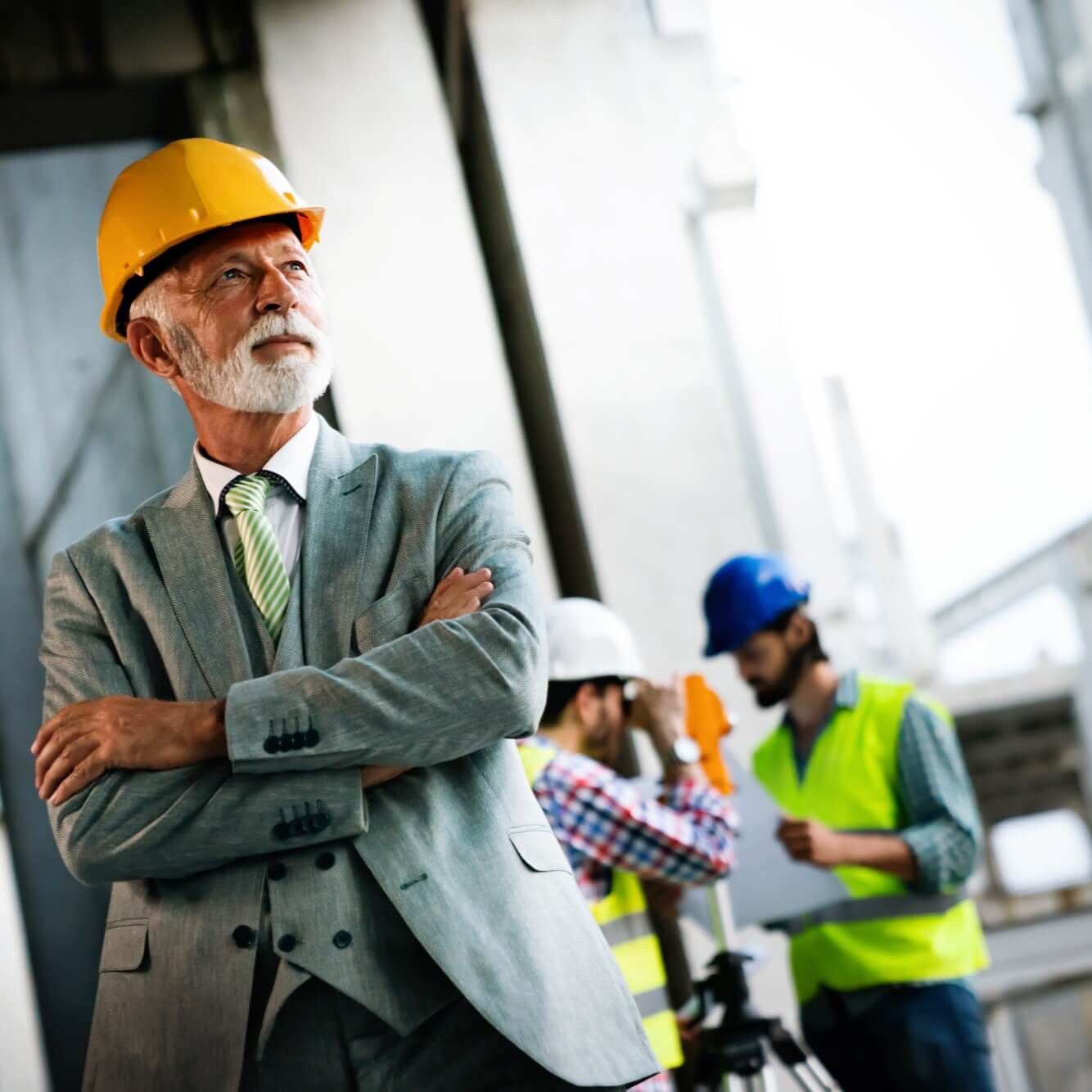 Stavebný tím: muž v obleku s prilbou v popredí, technici s plánom a meracím prístrojom v pozadí