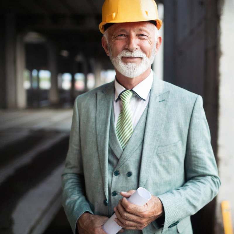 Stavebný dohľad: muž v zelenom obleku s prilbou drží plány na stavenisku medzi betónovými stĺpmi