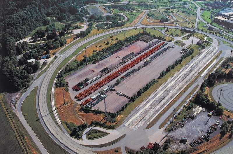 Letecký pohled na rozsáhlý testovací areál s různými typy drah, zaparkovanými vozy a okolními budovami