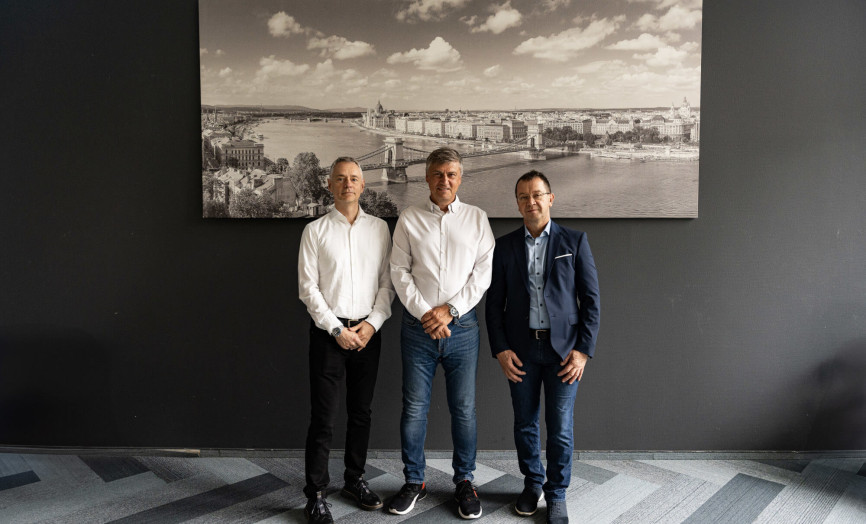 Three men in smart casual business attire standing in front of a large photograph of Budapest.