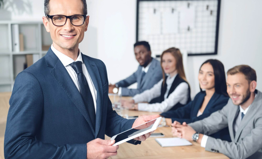 Usmívající se manažer v brýlích drží tablet v popředí, zatímco jeho tým sedí u jednacího stolu v pozadí.