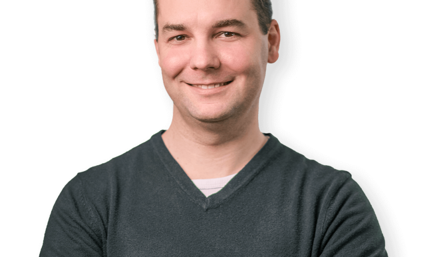 Studiový portrét usmívajícího se muže v tmavě zeleném svetru s výstřihem do V na neutrálním šedém pozadí; muž má krátké hnědé vlasy a působí profesionálním, přístupným dojmem.