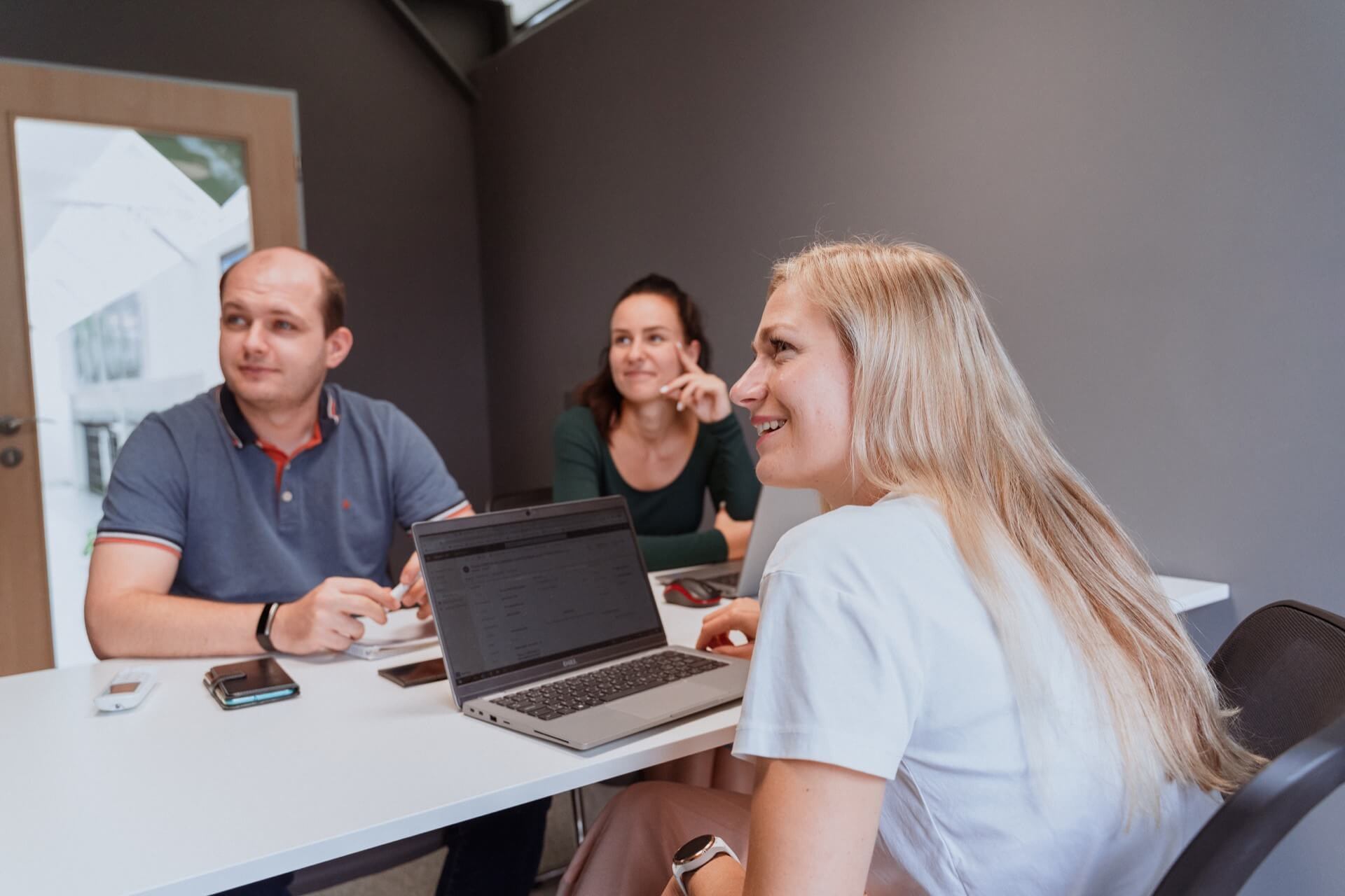 Tři lidé sedí kolem bílého stolu v moderní kanceláři, každý s notebookem, působí to jako neformální týmová diskuse nebo brainstorming