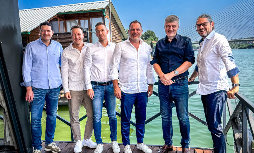 Six smiling men in casual business attire posing on the wooden deck of a houseboat; the background features a body of water, the opposite shore, and a modern cable-stayed bridge under a clear blue sky.