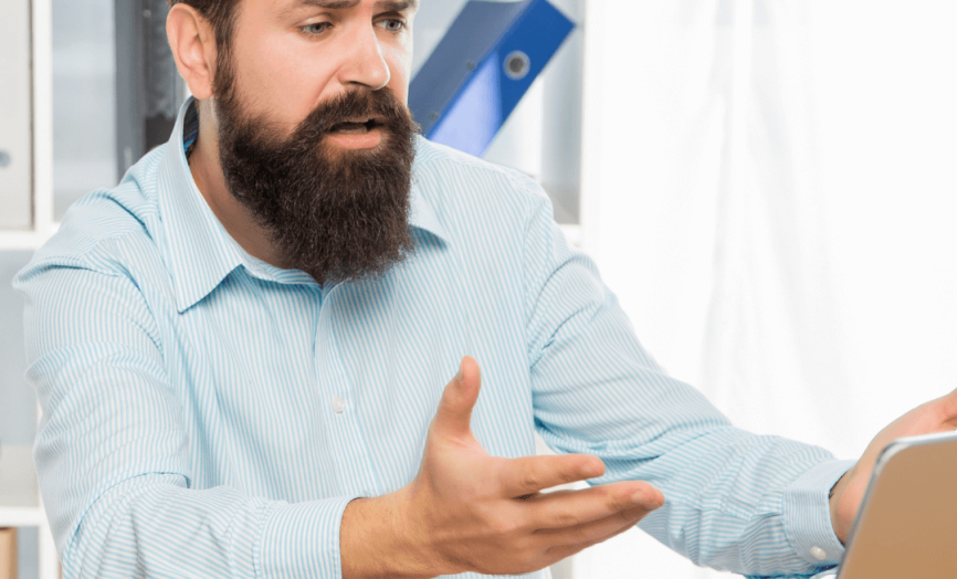 Rozrušený muž s plnovousem sedí v kanceláři u pracovního stolu a s nevěřícným výrazem a rozpaženýma rukama hledí na obrazovku svého notebooku.